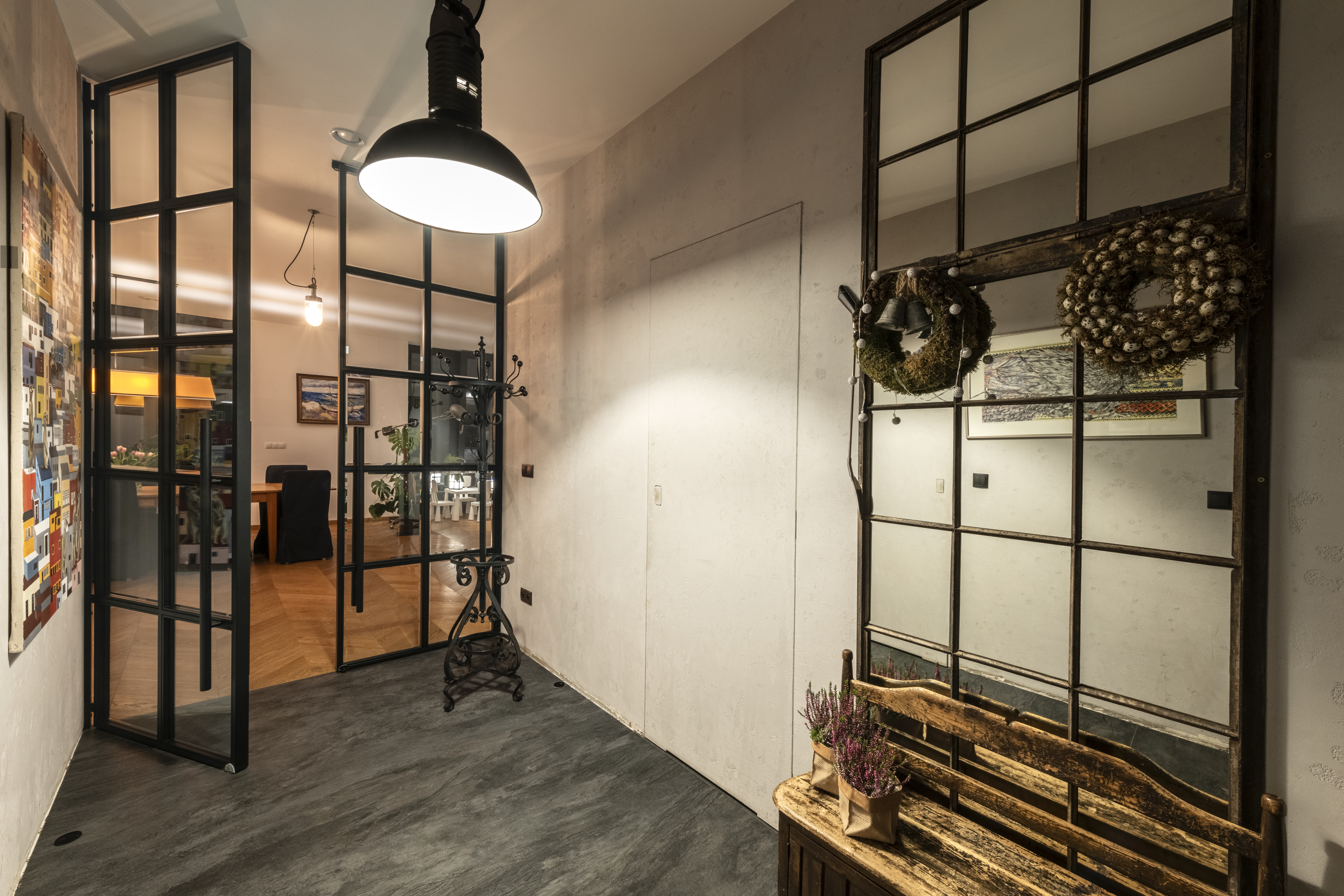 Stylowy hol wejściowy luksusowej willi, łączący industrialny design z rustykalnymi akcentami. Widoczna jest czarna szklana ścianka działowa, duża lampa wisząca w stylu industrialnym, drewniana ławka, ozdobne lustro w ramie okiennej z wiankami oraz...