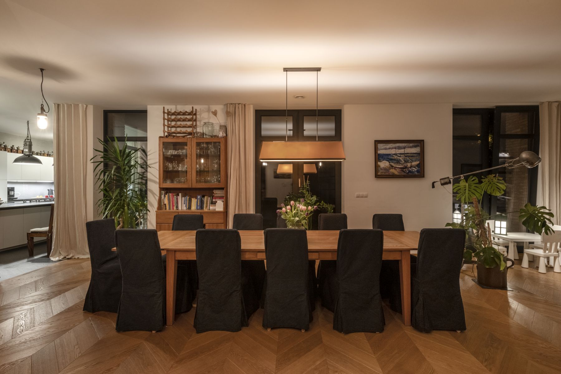 Przestronna, elegancka jadalnia z du偶ym drewnianym sto艂em na ponad 10 os贸b, czarnymi krzes艂ami, designersk膮 lamp膮 wisz膮c膮, pod艂og膮 w jode艂k臋 i widokiem na otwart膮 kuchni臋, tworz膮ca ciep艂膮 atmosfer臋.