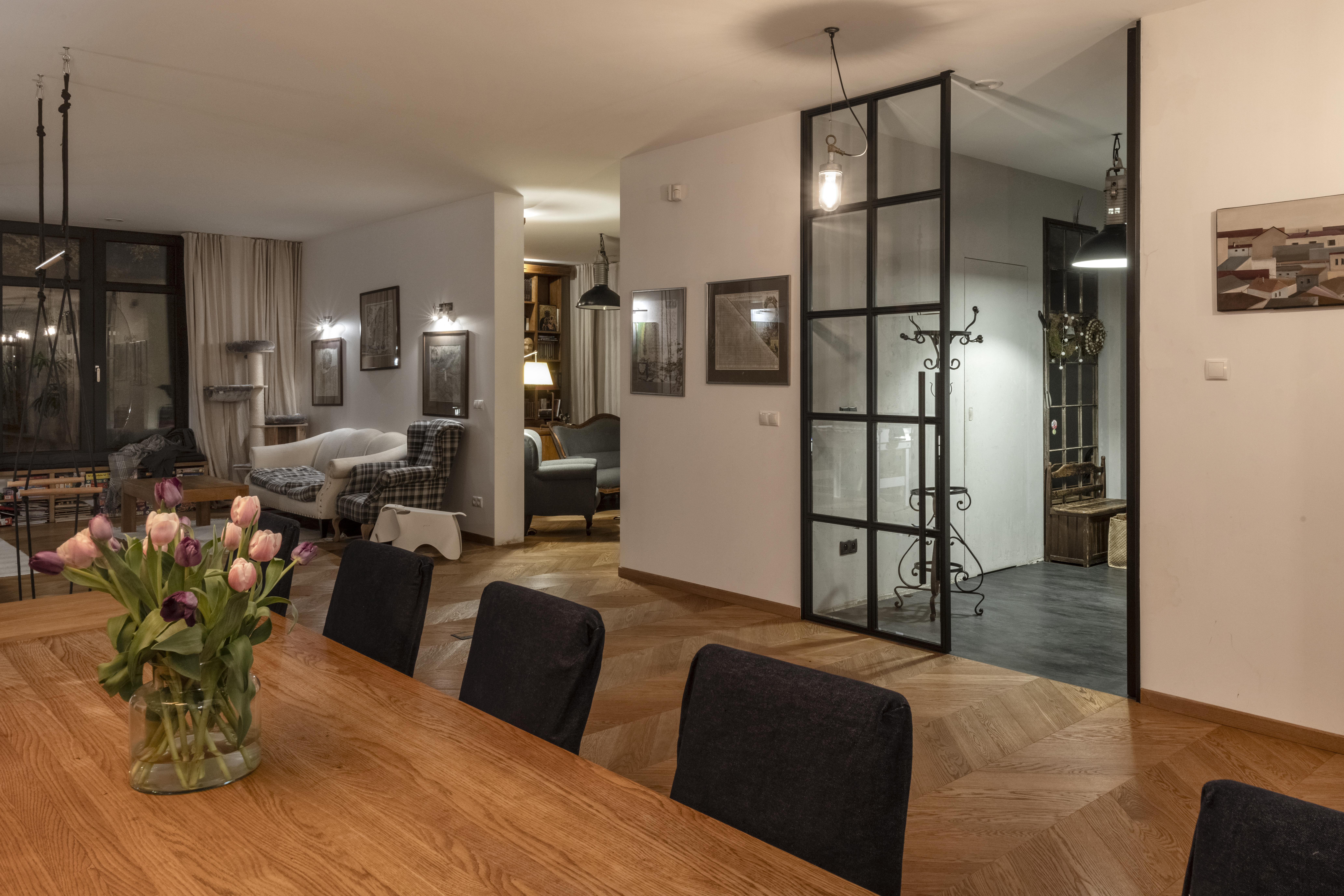 Elegancka, otwarta przestrze艅 艂膮cz膮ca jadalni臋 z salonem, z drewnianym sto艂em, wygodnymi krzes艂ami, sof膮, fotelami, ozdobn膮 艣ciank膮 ze szk艂a i metalu oraz pod艂og膮 w jode艂k臋. Ciep艂e o艣wietlenie tworzy luksusow膮 i przytuln膮 atmosfer臋.