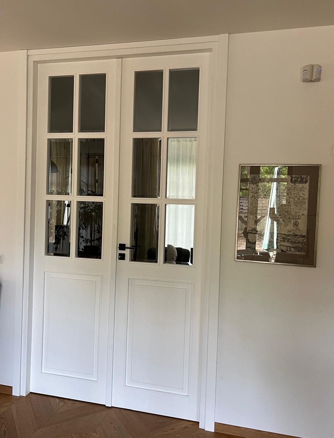 Białe drzwi francuskie z wieloma przeszkleniami, otwarte na jasny korytarz z jodełkową podłogą z drewna. Na białej ścianie obok wisi oprawiony obraz. Scena jest dobrze oświetlona naturalnym światłem, a drzwi mają ciemne klamki.