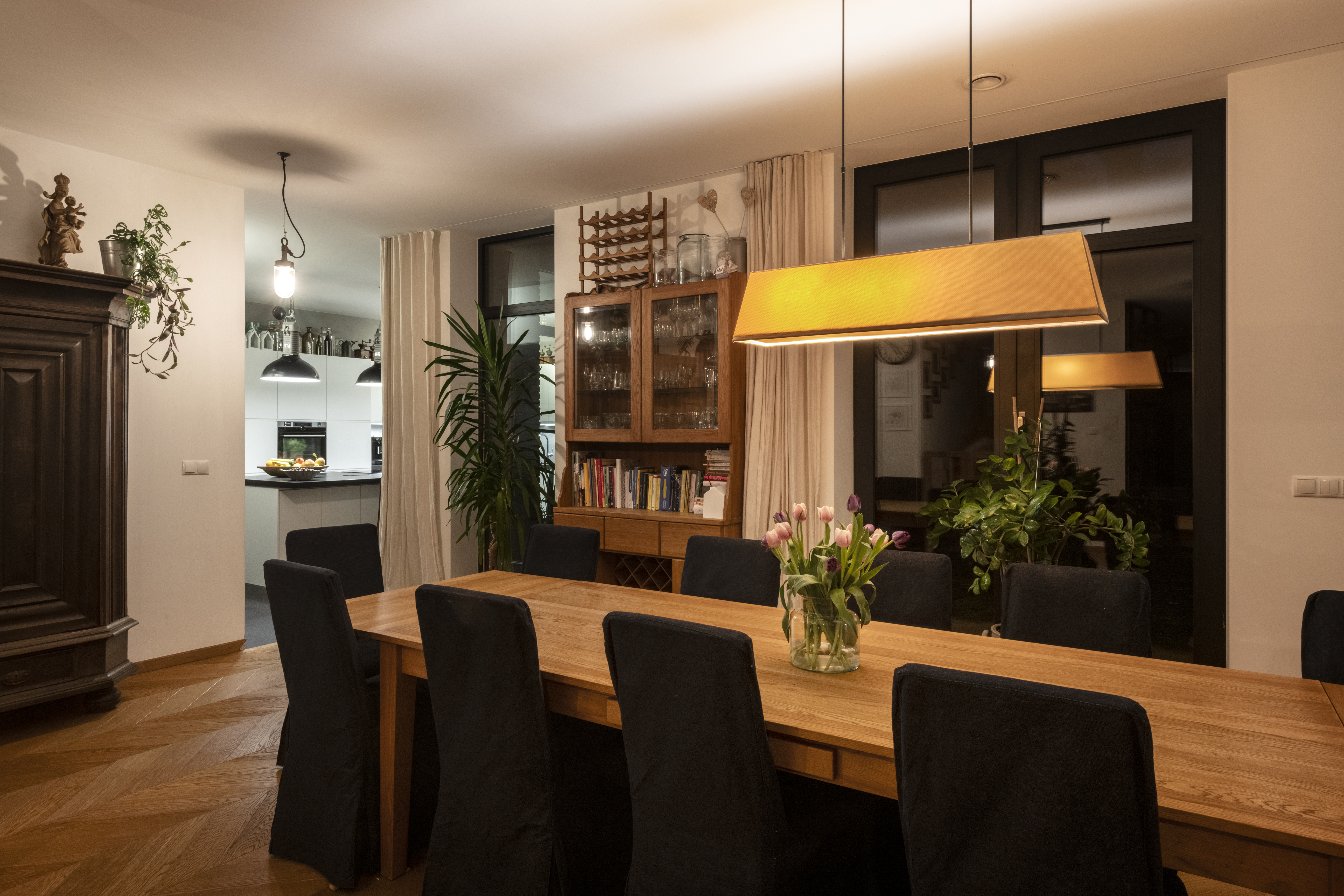 Elegancka jadalnia z du偶ym drewnianym sto艂em na osiem os贸b, wygodnymi krzes艂ami, nowoczesn膮 lamp膮 wisz膮c膮, przeszklon膮 witryn膮 z winem i zielonymi ro艣linami. W tle widoczna nowoczesna kuchnia. Idealna do wsp贸lnych posi艂k贸w.